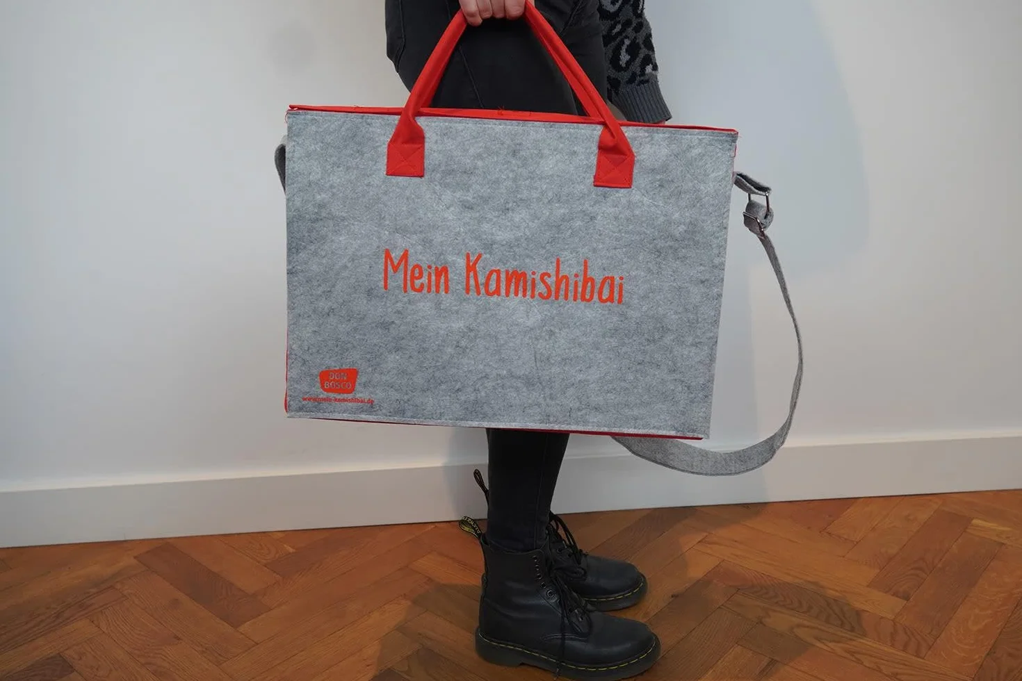 Tragetasche und Umhängetasche "Mein Kamishibai": Modell 2022. Grauer Filz, rote Seitenteile, verstärkter Boden, Klettverschluss, zwei Tragegriffe für die Hand, längenverstellbarer Schulterriemen